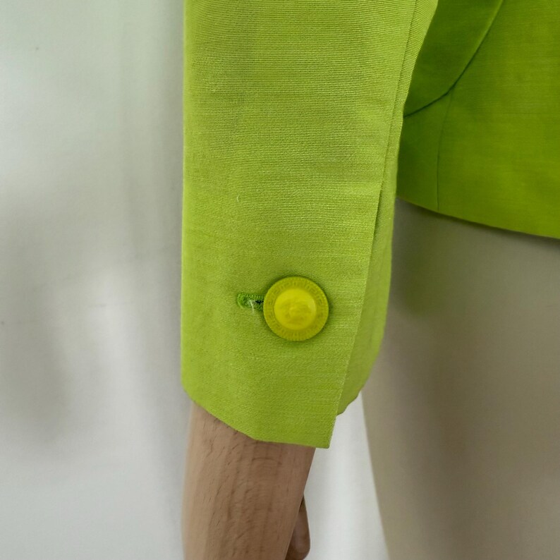 May include: Close-up of a lime green blazer sleeve with a matching button. The button is round and has a yellow center. The fabric has a subtle texture.