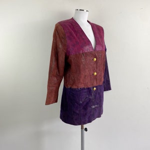 May include: A vintage leather jacket in a colour-block design. The jacket features a V-neck and gold buttons. The top section is a mix of burgundy and purple, while the bottom is a deep purple. The jacket is displayed on a mannequin.