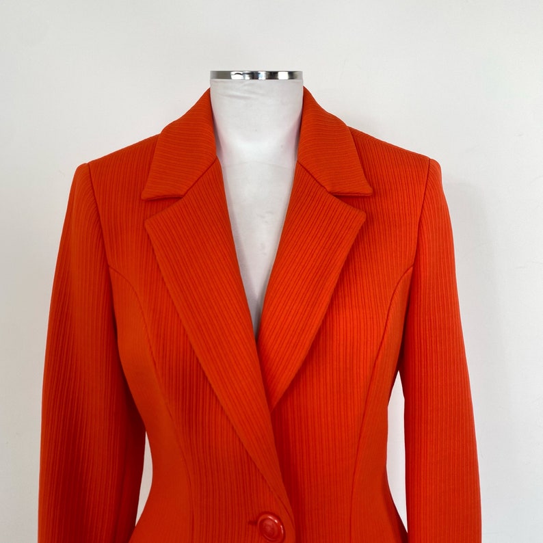 May include: A bright orange blazer with a single button closure and a ribbed texture. The blazer has a classic, tailored fit.