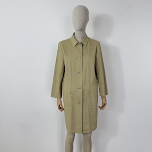 Puede incluir: Un abrigo beige con botones, cuello y dos bolsillos delanteros. El abrigo, a la altura de la rodilla, tiene mangas largas y se exhibe en un maniquí. El abrigo está hecho de una tela duradera. El abrigo es de color neutro.