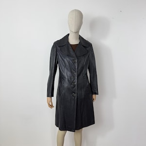 May include: A black leather coat with a button-up front, long sleeves, and a collared neckline. The coat is knee-length and appears to be vintage. The coat is displayed on a mannequin against a white background.