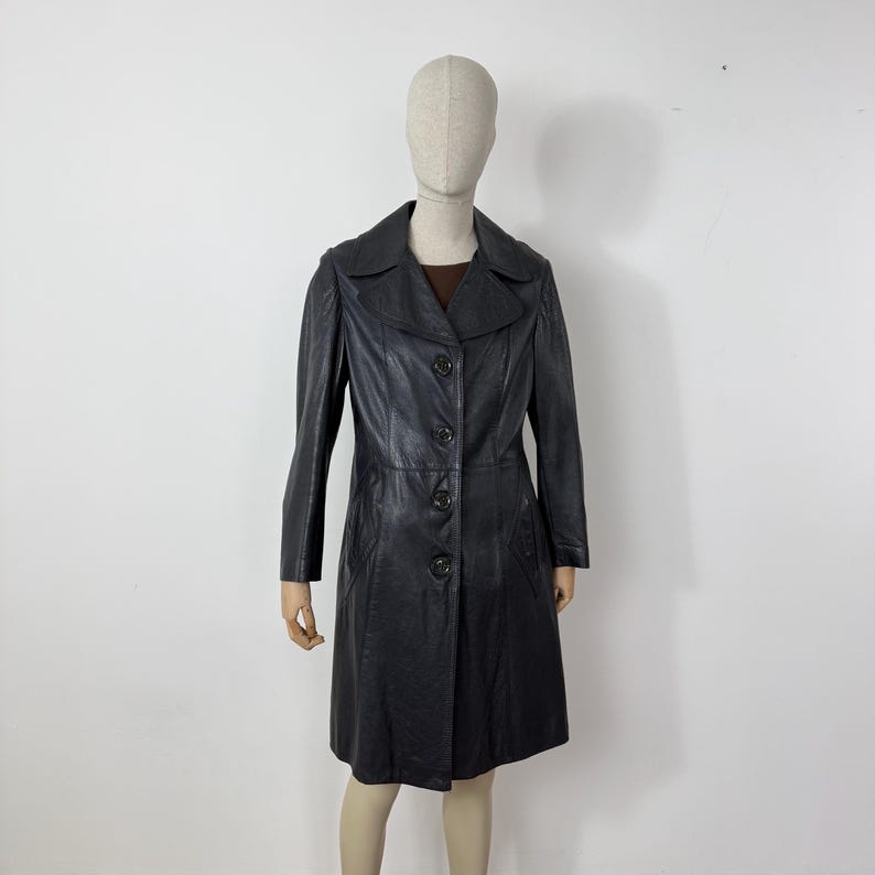 May include: A black leather coat with a button-up front, long sleeves, and a collared neckline. The coat is knee-length and appears to be vintage. The coat is displayed on a mannequin against a white background.