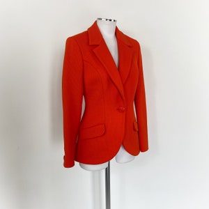 May include: A bright orange blazer with a single button closure and a notched collar. The blazer has a fitted silhouette and is made of a textured fabric.