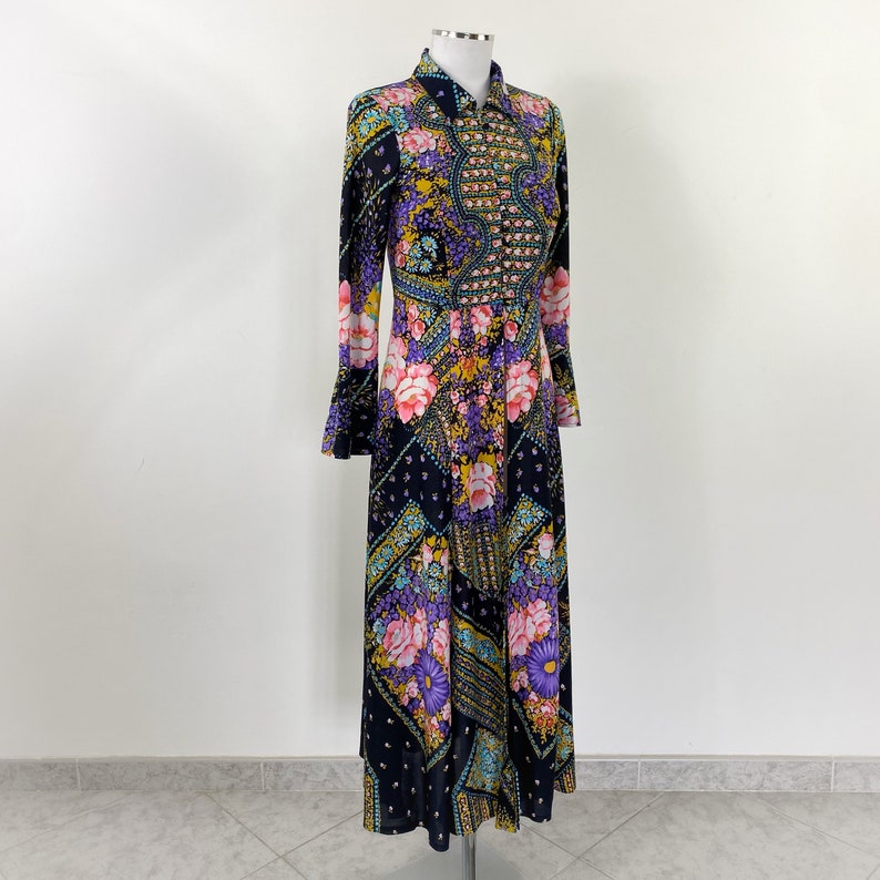 Puede incluir: Un vestido largo de manga larga con estampado floral en tonos rosa, morado, azul y amarillo sobre un fondo azul oscuro. El vestido tiene cuello y cierre de botones en la parte delantera.