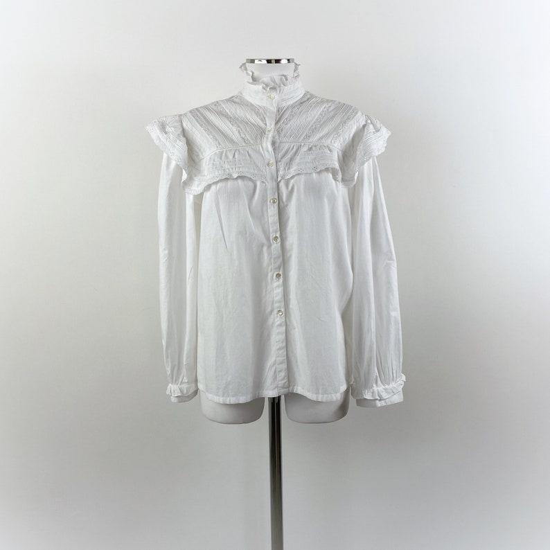 Puede incluir: Una camisa blanca abotonada con cuello alto con volantes y detalles de volantes en los hombros. La camisa de manga larga tiene una fila de botones en la parte delantera y se muestra en un maniqu&iacute;.