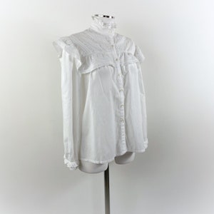 Puede incluir: Una camisa blanca abotonada con cuello alto con volantes y detalles de hombros con volantes. La camisa de manga larga tiene una fila de botones en la parte delantera y se muestra en un maniqu&iacute;.