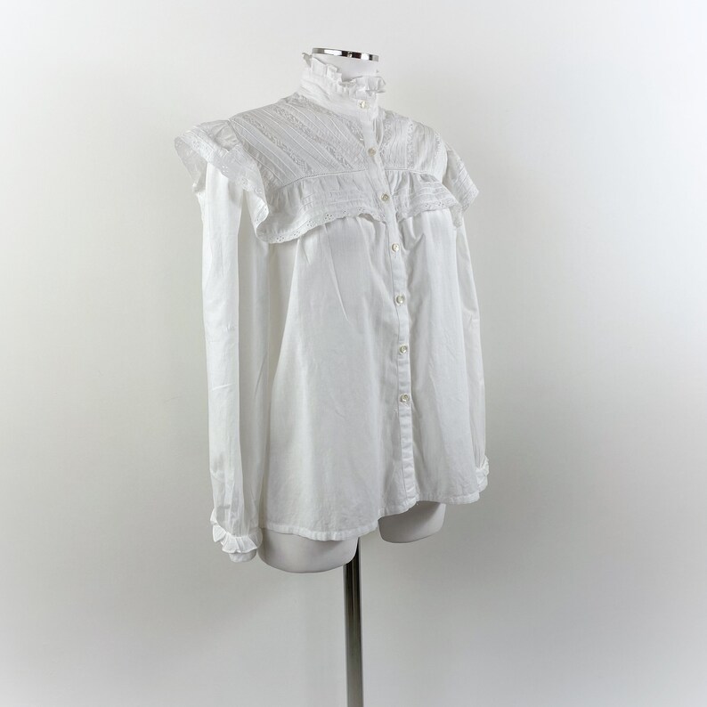Puede incluir: Una camisa blanca abotonada con cuello alto con volantes y detalles de hombros con volantes. La camisa de manga larga tiene una fila de botones en la parte delantera y se muestra en un maniqu&iacute;.