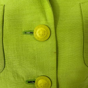 May include: Close-up of a lime green blazer with two large, round, yellow buttons. The buttons feature a detailed design with a central emblem. The blazer has a tailored fit and visible stitching.