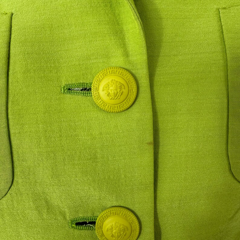 May include: Close-up of a lime green blazer with two large, round, yellow buttons. The buttons feature a detailed design with a central emblem. The blazer has a tailored fit and visible stitching.