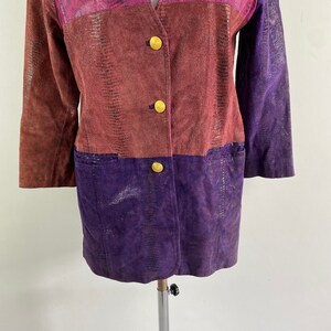 May include: A colour-blocked jacket with a reptile-like texture. The jacket features a V-neck and three gold buttons. The colour scheme includes shades of brown, purple, and pink. The jacket has two pockets.