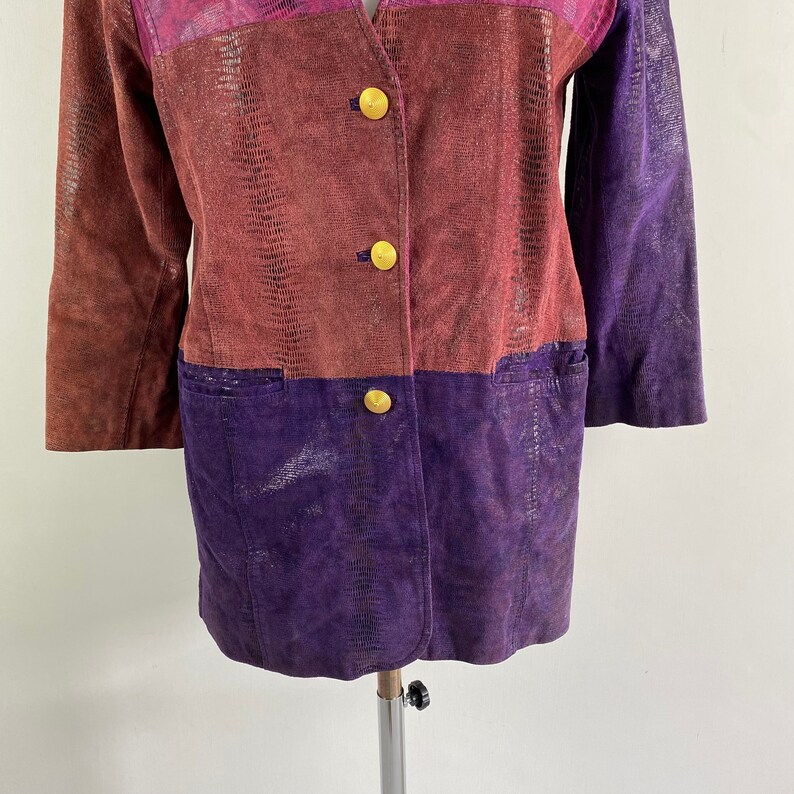May include: A colour-blocked jacket with a reptile-like texture. The jacket features a V-neck and three gold buttons. The colour scheme includes shades of brown, purple, and pink. The jacket has two pockets.