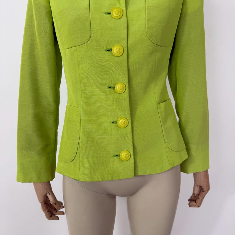 May include: A lime green blazer with a tailored fit. The jacket features a notched collar, two front pockets, and bright yellow buttons. The long sleeves and structured design create a classic silhouette. The fabric has a subtle texture.