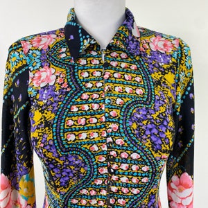 Puede incluir: Una camisa de manga larga con fondo negro y un estampado paisley colorido. La camisa tiene cuello y cierre de botones en la parte delantera.