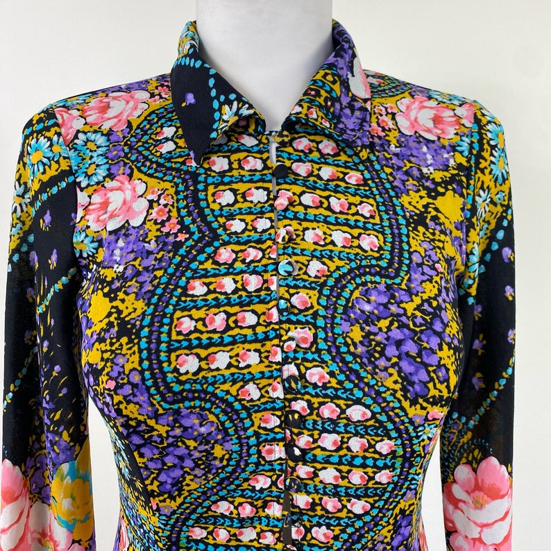 Puede incluir: Una camisa de manga larga con fondo negro y un estampado paisley colorido. La camisa tiene cuello y cierre de botones en la parte delantera.