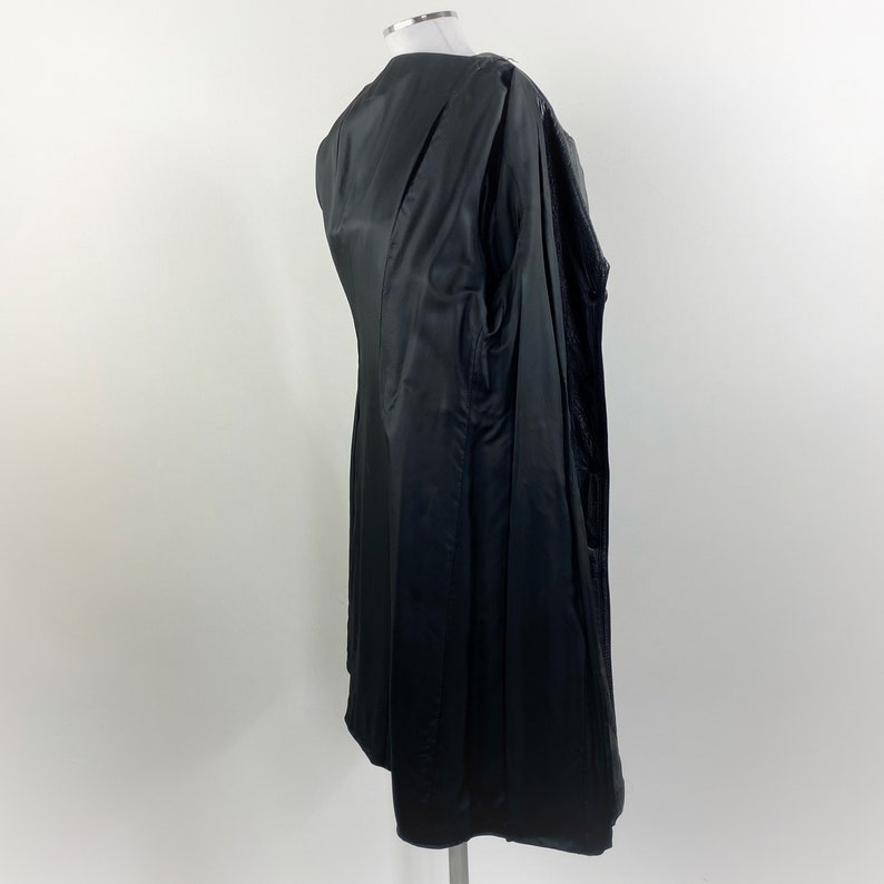 May include: A black coat with a satin-like sheen, displayed on a mannequin. The coat is long, with a tailored fit and a visible interior lining. The garment is designed with a classic silhouette.