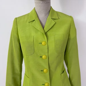 May include: A bright green blazer with a classic design. The blazer features a notched collar, two front pockets, and four matching yellow buttons. The fabric has a subtle sheen, giving it a polished look. The sleeves are long and tailored.