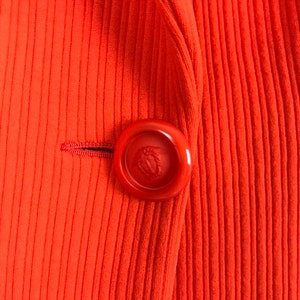 May include: A close-up of a bright orange ribbed blazer with a large red button featuring a gold emblem.