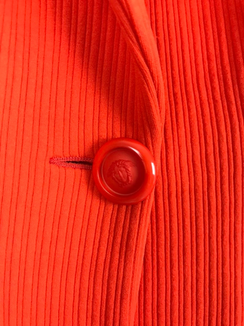 May include: A close-up of a bright orange ribbed blazer with a large red button featuring a gold emblem.