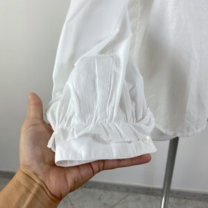 Puede incluir: Primer plano de una blusa blanca de manga larga. La manga presenta un pu&ntilde;o con volantes y un cierre abotonado. La tela parece ligera y ligeramente texturizada.