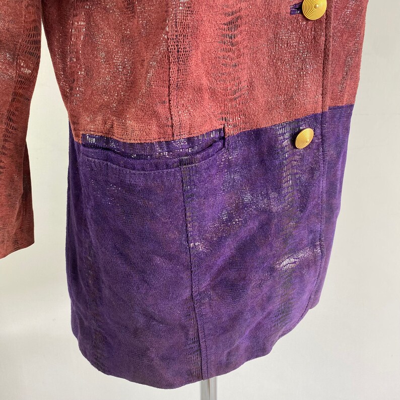May include: A two-tone blazer with a reptile-like texture. The upper section is a muted reddish-brown, and the lower section is a deep purple. Gold-coloured buttons fasten the front. The blazer features a pocket on the lower half.