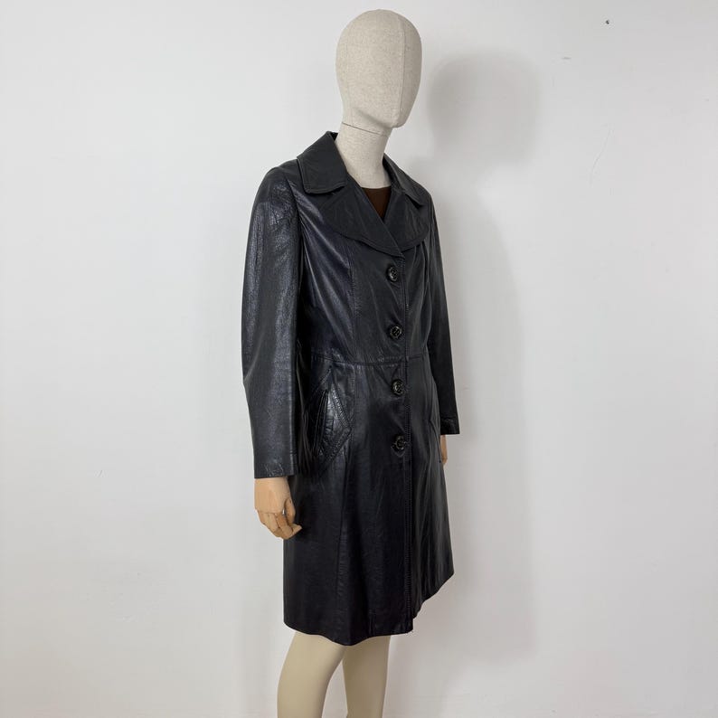May include: A black leather coat with a classic design. The coat features a collared neckline, button-down closure, and long sleeves. The coat is displayed on a mannequin against a white background. The coat appears to be knee-length.