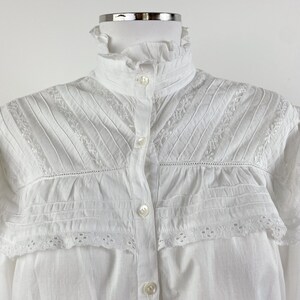 Puede incluir: Camisa blanca abotonada con cuello alto con volantes y delicados detalles de encaje y pliegues. La camisa presenta una serie de peque&ntilde;os botones en la parte delantera y bordados intrincados a lo largo del canes&uacute; y los pu&ntilde;os.