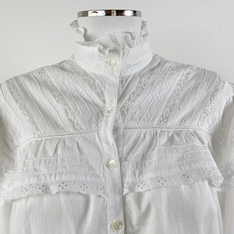 Puede incluir: Camisa blanca abotonada con cuello alto con volantes y delicados detalles de encaje y pliegues. La camisa presenta una serie de peque&ntilde;os botones en la parte delantera y bordados intrincados a lo largo del canes&uacute; y los pu&ntilde;os.