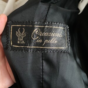 May include: Black interior of a jacket with a leather label. The label has gold lettering and a stylized antelope emblem. The text on the label reads "Creazioni in pelle".