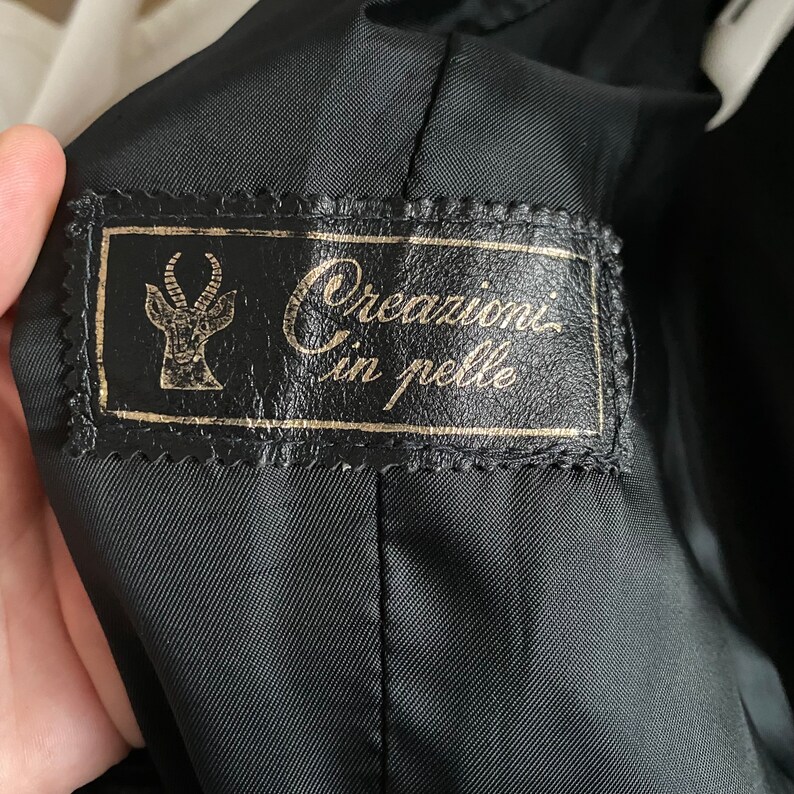 May include: Black interior of a jacket with a leather label. The label has gold lettering and a stylized antelope emblem. The text on the label reads "Creazioni in pelle".