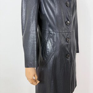 May include: A black leather coat with a button-up front and a tailored fit. The coat features a notched collar, a diamond-shaped pocket, and a knee-length hem. The coat is made of a smooth, shiny leather material.