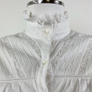 Puede incluir: Una blusa blanca con cuello alto con volantes y cierre de botones. La blusa presenta pliegues verticales y delicados detalles florales bordados a lo largo del canes&uacute; y las mangas. La tela parece ser de algod&oacute;n o lino ligero.