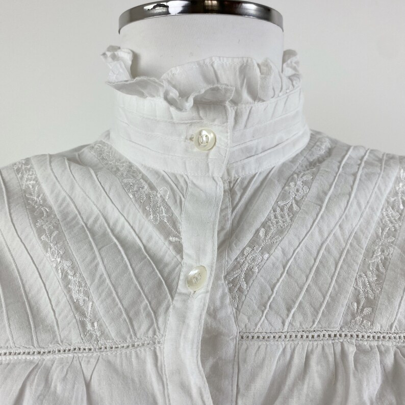 Puede incluir: Una blusa blanca con cuello alto con volantes y cierre de botones. La blusa presenta pliegues verticales y delicados detalles florales bordados a lo largo del canes&uacute; y las mangas. La tela parece ser de algod&oacute;n o lino ligero.