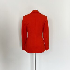 May include: A bright orange blazer with a classic cut and a single-breasted closure. The blazer is made of a textured fabric and has a slightly fitted silhouette.