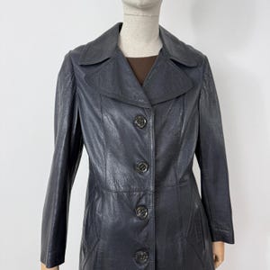 May include: A vintage, dark navy blue leather coat with a classic design. The coat features a notched collar, long sleeves, and a button-down front. The coat is approximately mid-length and has a tailored fit, suitable for various occasions.