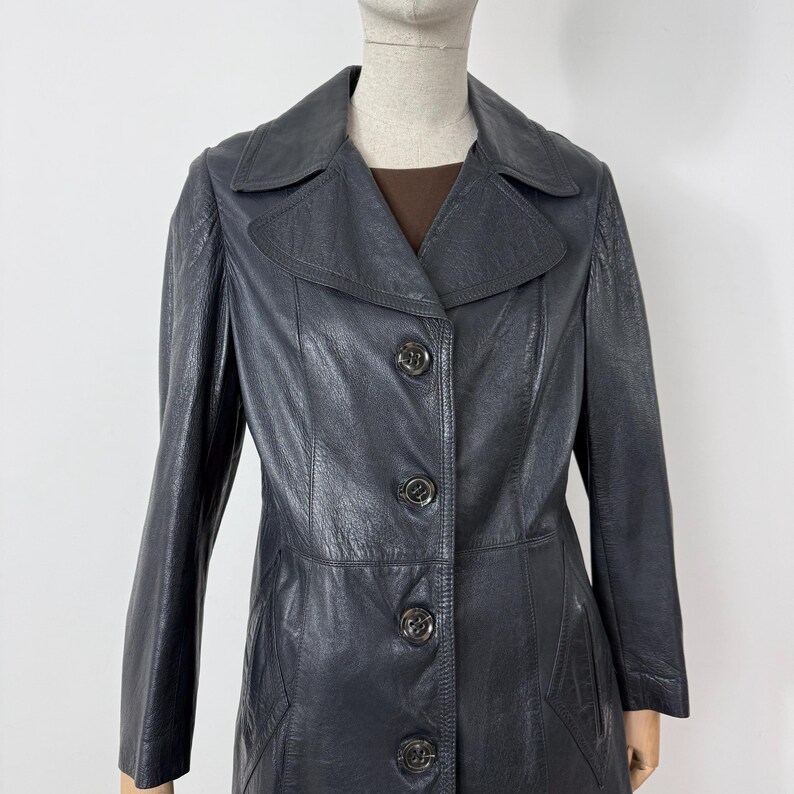 May include: A vintage, dark navy blue leather coat with a classic design. The coat features a notched collar, long sleeves, and a button-down front. The coat is approximately mid-length and has a tailored fit, suitable for various occasions.