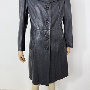 May include: A black leather coat with a button-up front, long sleeves, and a collared neckline. The coat has a tailored fit and appears to be knee-length. The buttons are a dark, possibly antique, metal. The coat is displayed on a mannequin.