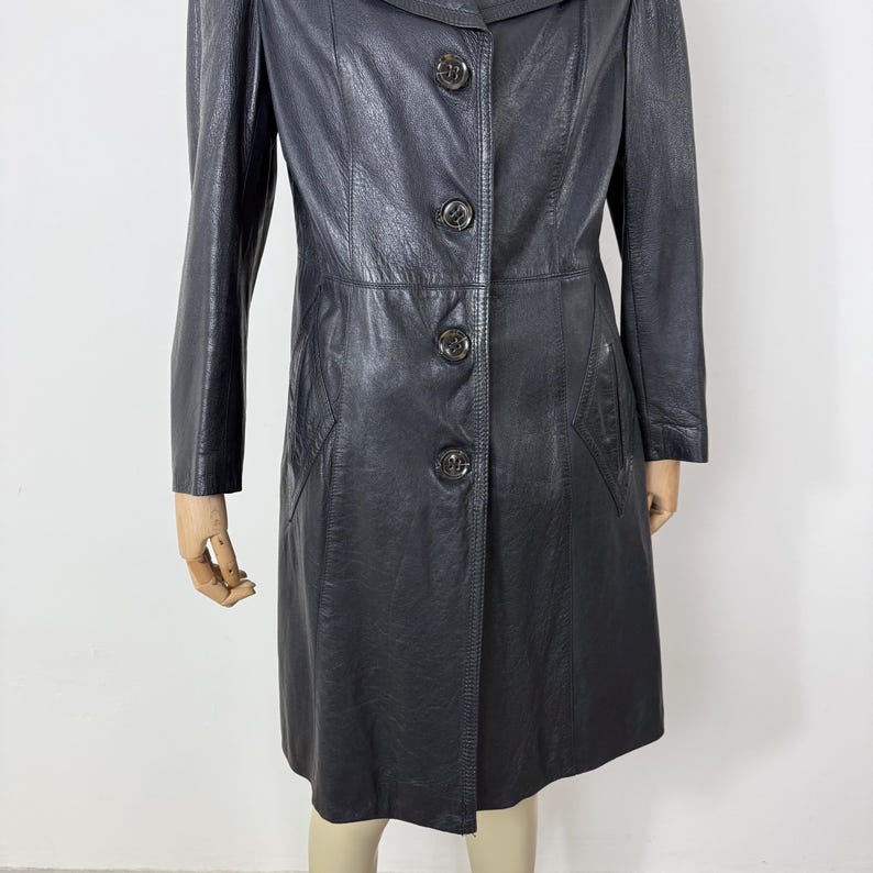 May include: A black leather coat with a button-up front, long sleeves, and a collared neckline. The coat has a tailored fit and appears to be knee-length. The buttons are a dark, possibly antique, metal. The coat is displayed on a mannequin.