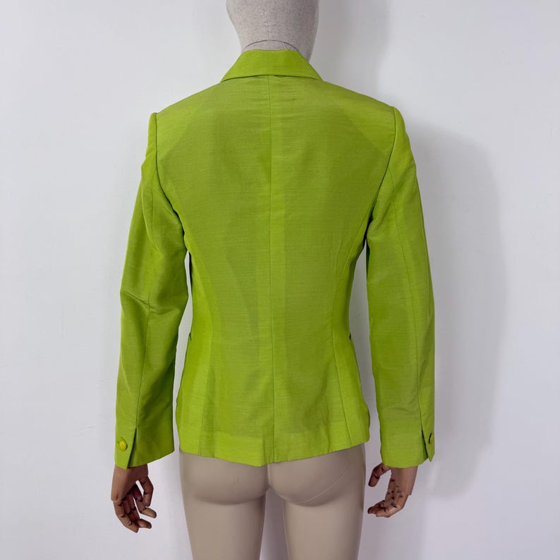 May include: A lime green blazer with a two-button closure and long sleeves. The blazer is displayed on a mannequin, showcasing the back of the garment. The fabric has a subtle sheen, suggesting a silk or similar material. The blazer has a classic tailored design.