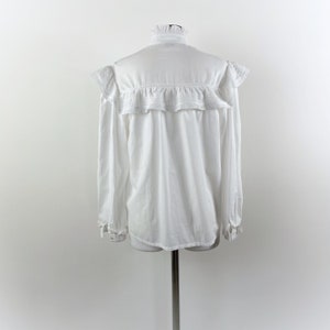 Puede incluir: Una blusa blanca de manga larga con cuello alto con volantes y detalles de volantes en los hombros y el pecho. La blusa se exhibe en un maniqu&iacute; sobre un fondo blanco liso. Los pu&ntilde;os tambi&eacute;n tienen detalles de volantes.