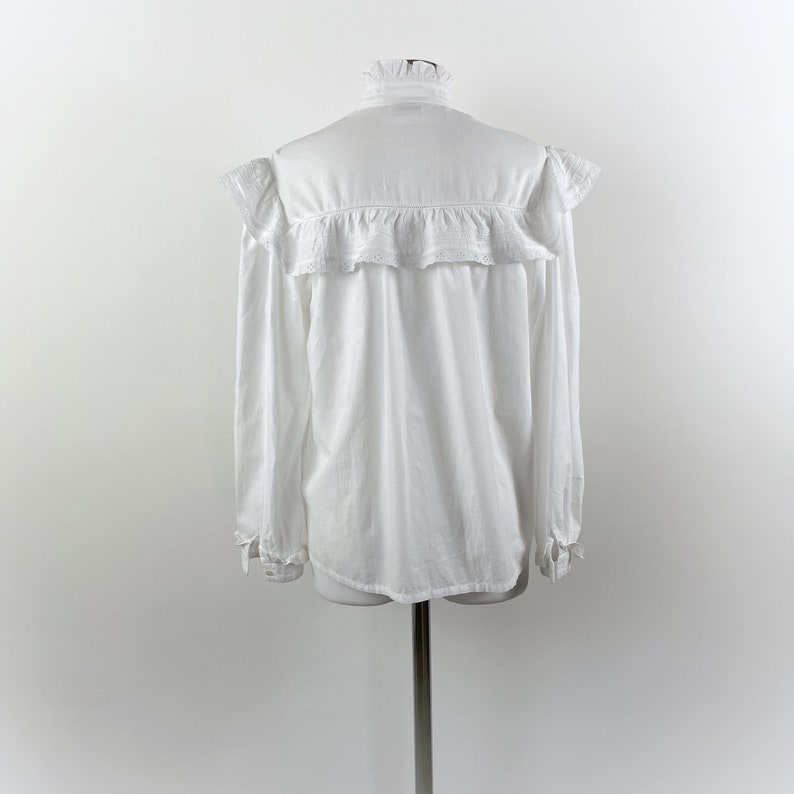 Puede incluir: Una blusa blanca de manga larga con cuello alto con volantes y detalles de volantes en los hombros y el pecho. La blusa se exhibe en un maniqu&iacute; sobre un fondo blanco liso. Los pu&ntilde;os tambi&eacute;n tienen detalles de volantes.