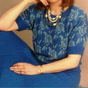 May include: A woman wearing a blue floral knit sweater and a matching skirt. She is wearing a multi-strand necklace with white and gold beads.