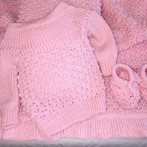May include: A pink knitted baby sweater, hat, and booties set. The sweater has a loose fit and a wide neckline. The hat and booties are both made with a textured stitch.