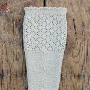 Op de afbeelding: Ivoorwitte gebreide beenwarmers met een ruitpatroon op het bovenste gedeelte en een geschulpte rand. De beenwarmers zijn gemaakt van een zacht, getextureerd garen en worden gepresenteerd op een rustiek houten oppervlak.