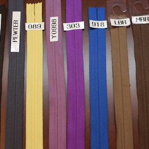 May include: A selection of ten zipper tapes in various colors, including black, charcoal, pewter, yellow, purple, blue, brown, and dark brown. The zipper tapes are labeled with their corresponding color codes: BK, CHARCOAL, PEWTER, 089, Y0998, 303, 918, LBR, MBR, and DBR.