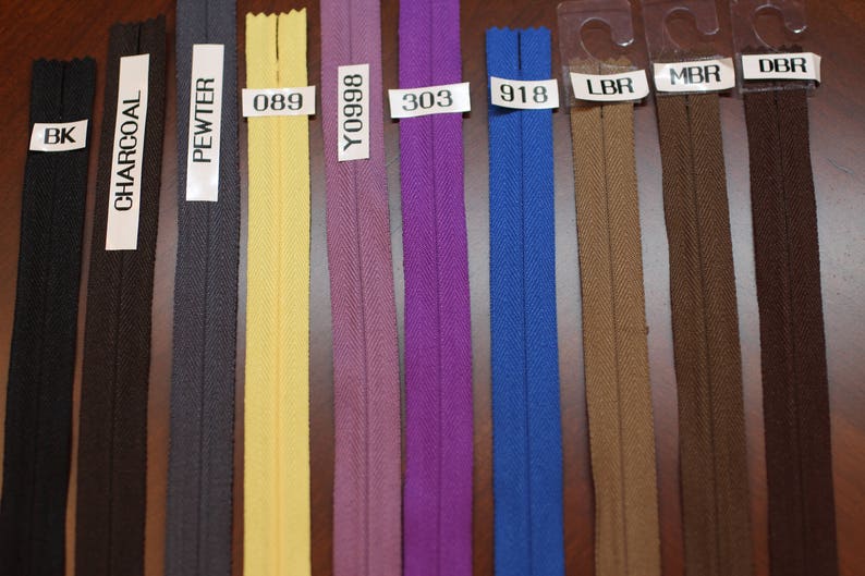 May include: A selection of ten zipper tapes in various colors, including black, charcoal, pewter, yellow, purple, blue, brown, and dark brown. The zipper tapes are labeled with their corresponding color codes: BK, CHARCOAL, PEWTER, 089, Y0998, 303, 918, LBR, MBR, and DBR.