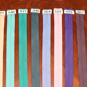 May include: A collection of nine long, narrow fabric zippers in various colors including yellow, peach, turquoise, teal, dark blue, light blue, pink, purple, and dark purple. Each zipper features a white plastic tag with a black number code.