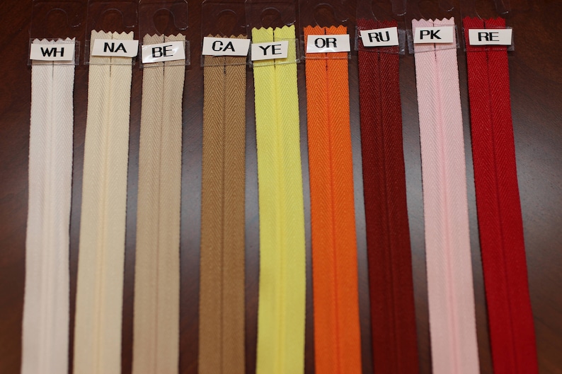 May include: A selection of zipper tapes in various colors, including white, beige, brown, yellow, orange, red, pink, and red. The zipper tapes are labeled with letters, including WH, NA, BE, CA, YE, OR, RU, PK, and RE.