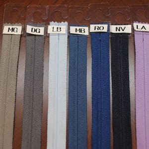 May include: A selection of ten zipper tapes in various colors, including beige, gray, light blue, dark blue, navy, black, lavender, and purple. The zipper tapes are displayed on a brown surface with labels indicating the color names.