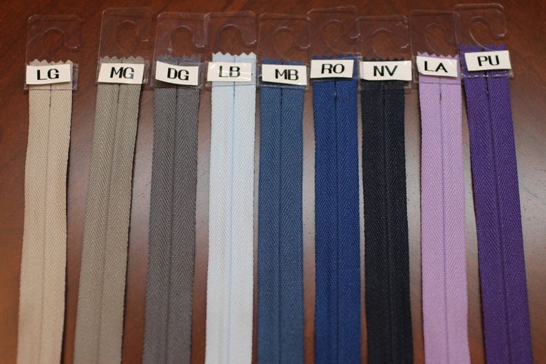 May include: A selection of ten zipper tapes in various colors, including beige, gray, light blue, dark blue, navy, black, lavender, and purple. The zipper tapes are displayed on a brown surface with labels indicating the color names.