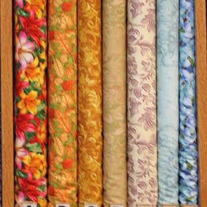 May include: Nine fabric rolls with different patterns, labeled A through G. The fabric rolls are arranged horizontally on a wooden shelf. The fabric patterns include floral, leaf, and geometric designs in various colors.